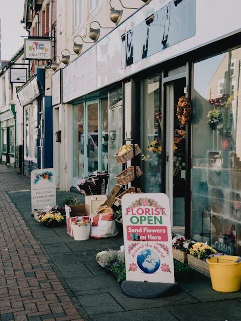 Exterior view of a flower shop on Barkingside High Street with large glass windows displaying floral arrangements. Outside the shop, there are buckets and baskets filled with fresh, colorful flowers s
