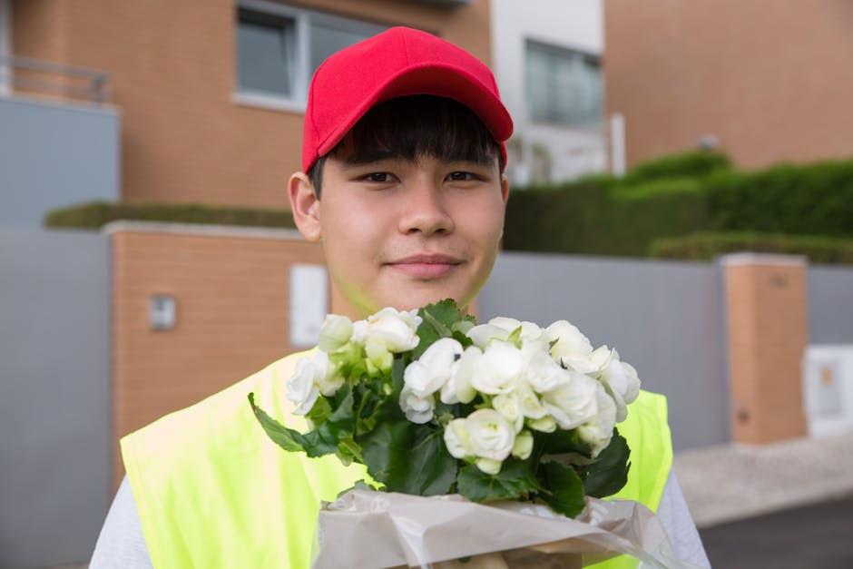 A young male delivery person wearing a red cap and a high-visibility yellow vest holds a bouquet of fresh white roses wrapped in white paper. He stands outdoors in front of modern residential building