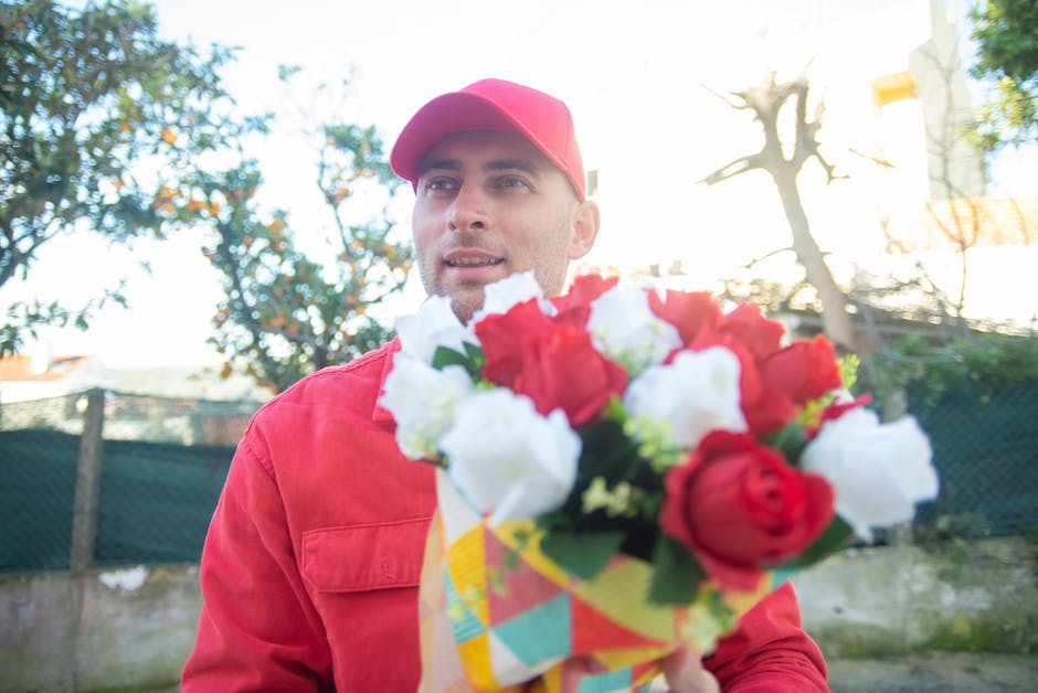 A delivery person dressed in a red cap and matching red jacket holds a vibrant bouquet of fresh flowers in front of him, with a background of trees and sunlight filtering through the leaves. The bouqu