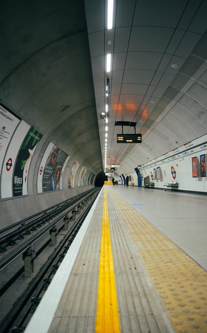 The image depicts an underground train station with a curved concrete ceiling and tiled walls. The platform features a tactile yellow safety line running parallel to the train tracks, with a textured