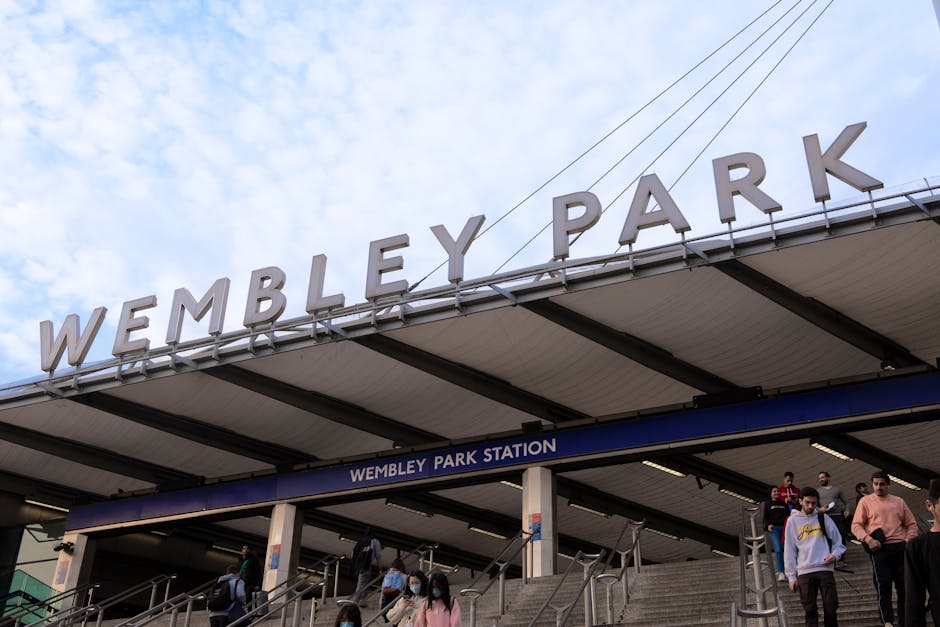 Wembley Park Station entrance with large white signage spelling 'WEMBLEY PARK' mounted on the roof, featuring a blue and white sign indicating 'WEMBLEY PARK STATION'. Several commuters, some wearing f