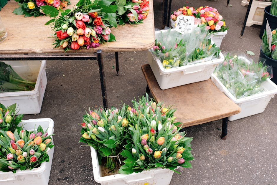 Vivid tulip bouquets with pink, yellow, white, and red blooms, arranged with green foliage, are displayed in white plastic containers and on a wooden table at a flower market. Some tulips are wrapped 